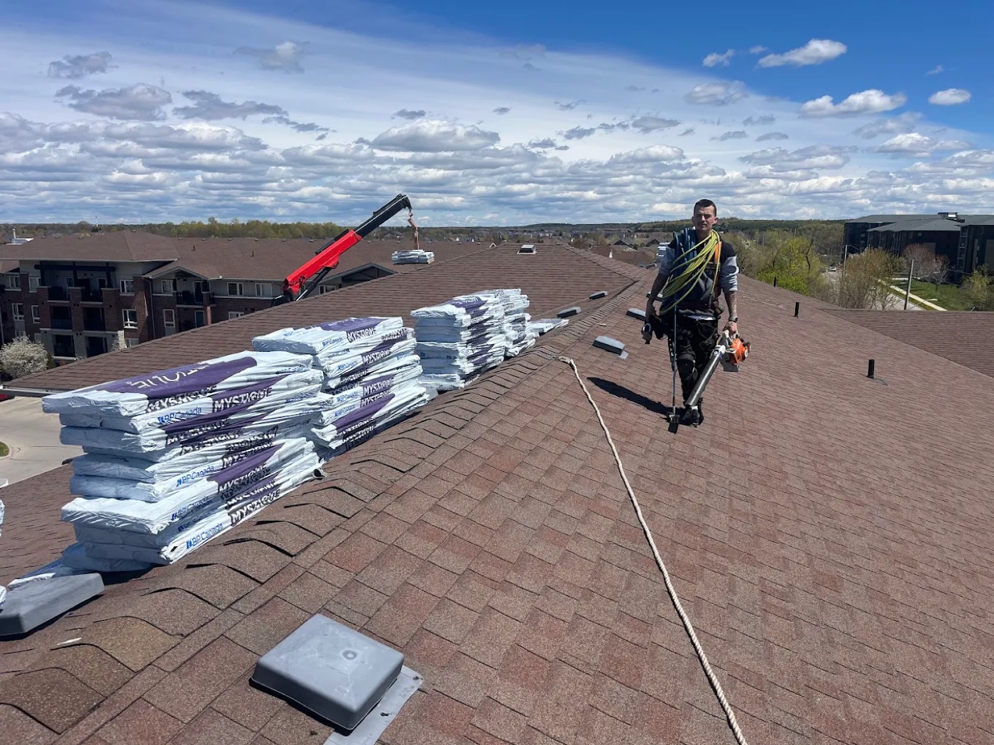 Roofer harnessed and walking a steep roof ridge with bundled shingles staged nearby, aerial view